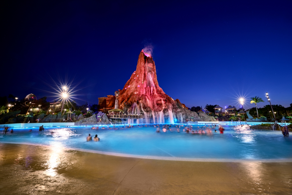 A glowing pool at night with people swimming, a towering and glowing red volcano in the background.