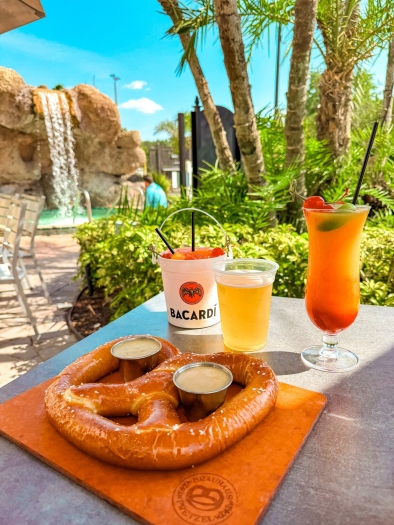 A large soft pretzel on a wooden board set on a table with a beer and two tropical cocktails, surrounded by tropical greenery with a waterfall in the background.