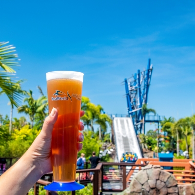 A tall pint glass of beer held against a blue sky with a raft water ride in the background.