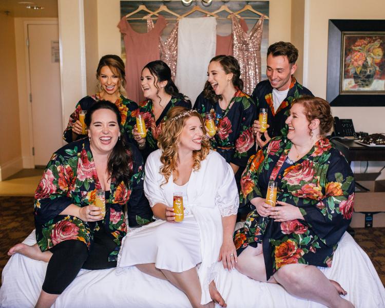 A bridal party in navy floral robes surround a bride sitting on a bed with dresses hanging in the background. 