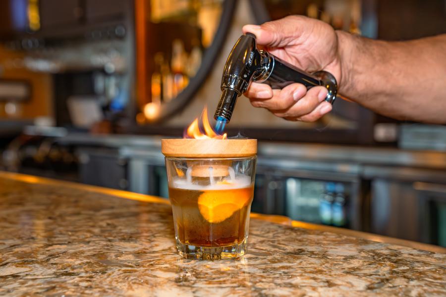 A hand with a culinary torch lights a smoker on top of a cocktail at Jack's Place Lobby Bar.