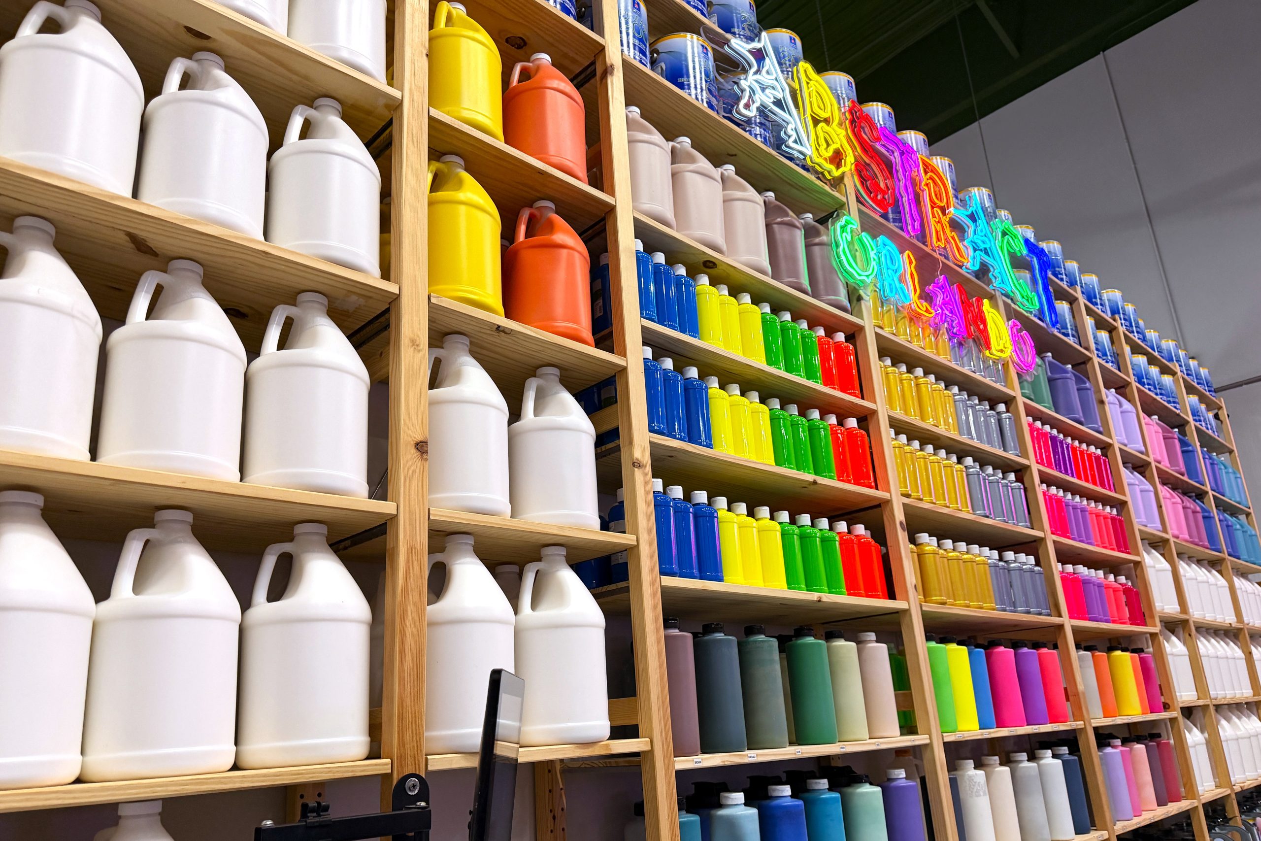 Tall shelves filled with brightly colored paint and a neon sign that reads Abstract Orlando.
