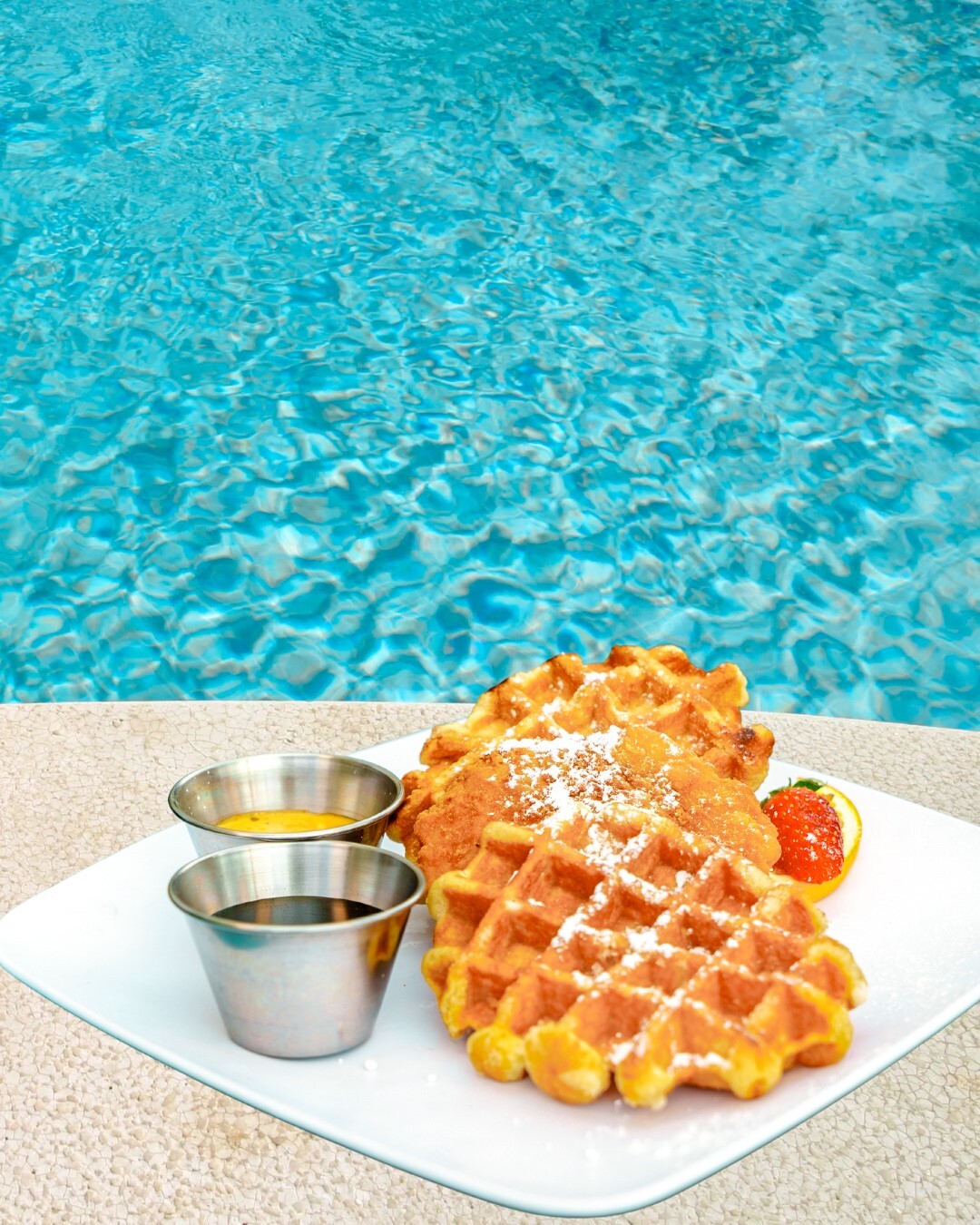 Two stacked waffles with syrup sit on a white plate beside sparkling pool water.