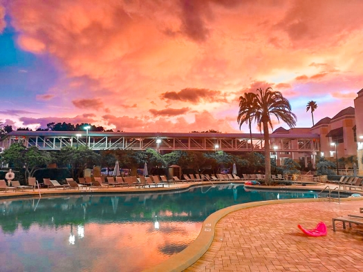 A large swimming pool at twilight with pink and orange clouds and the OCCC skybridge crossing across the horizon at Rosen Plaza.