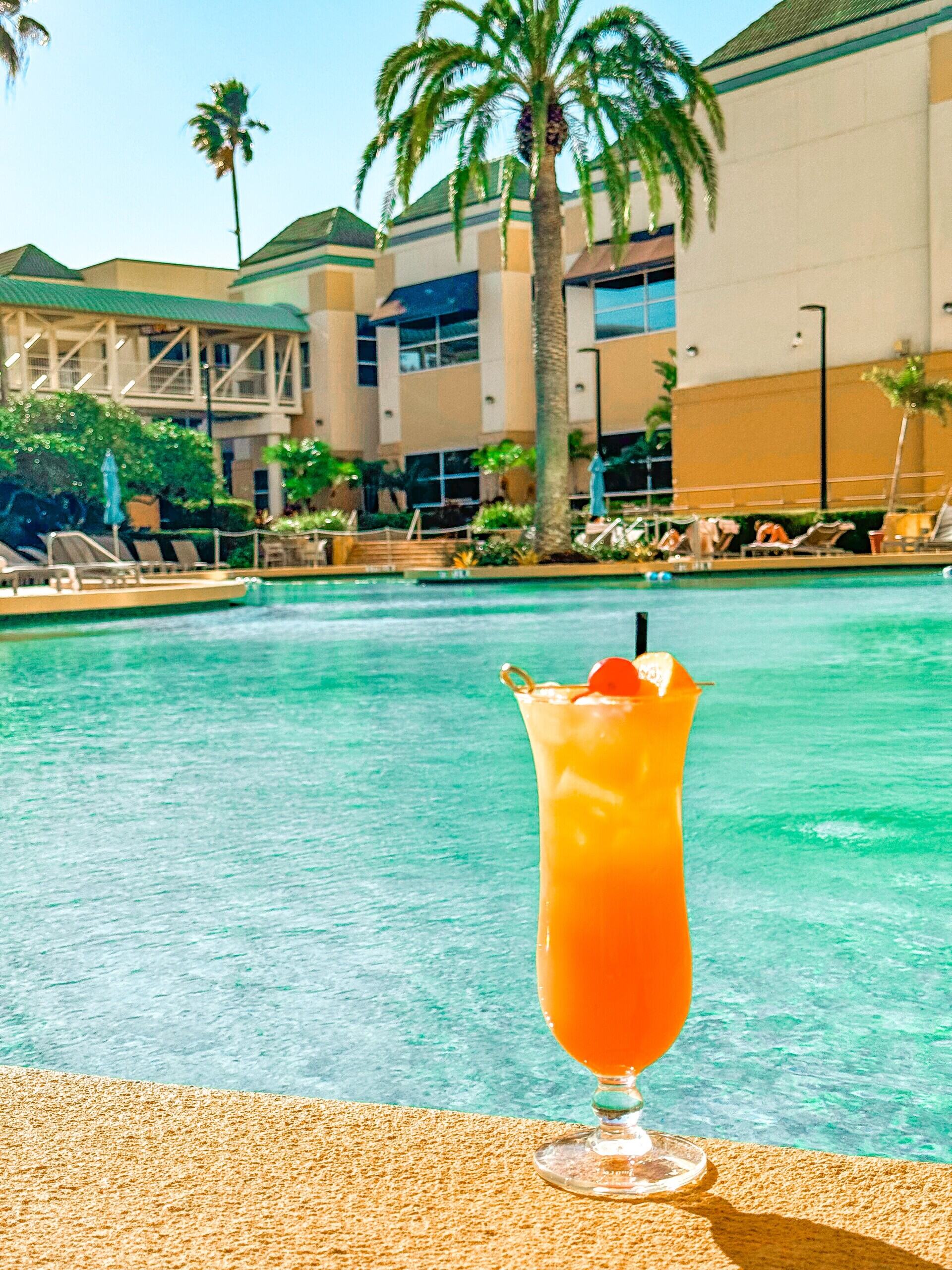An orange cocktail in a hurricane glass sits in front of a beautiful swimming pool and hotel.