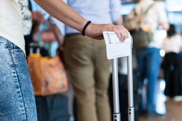 A close-up of a man's hand on a suitcase handle holding a ticket and standing in an airport security line.