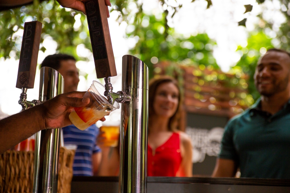 A bartender's hands pour a beer from a tap in front of an outdoor bar with guests.