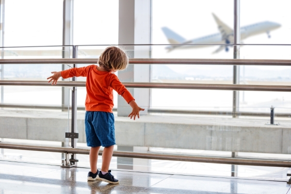 A boy in a red shirt stands in front of an airport window looking at an airplane with his arms outstretched.