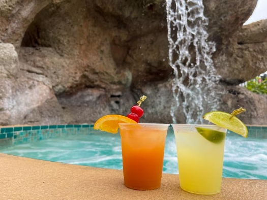 An orange and yellow cocktail sitting on the deck of a pool with a waterfall in the background.