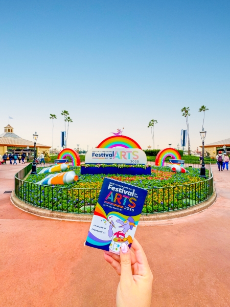 A hand holding a ticket in front of the entrance to the EPCOT International Festival of the Arts.
