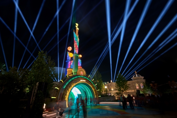 An arched entryway lit colorfully at night against a dark sky with rays of blue light shooting into the sky.