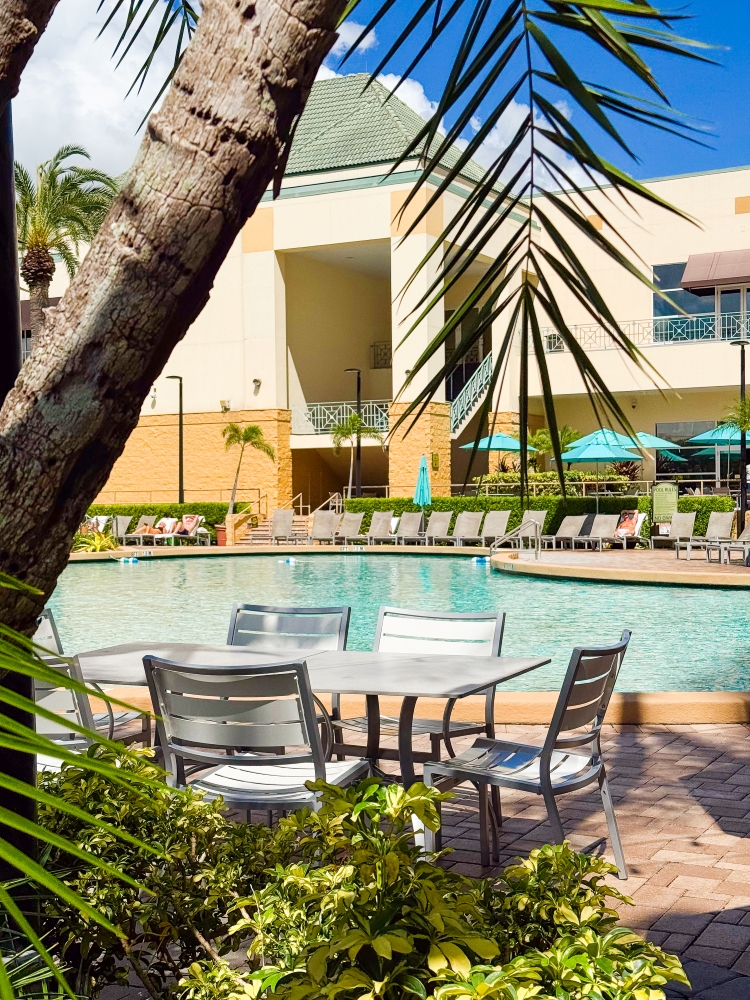 A table and chairs sit on a pool deck beneath palm branches, next to a large pool and a building.