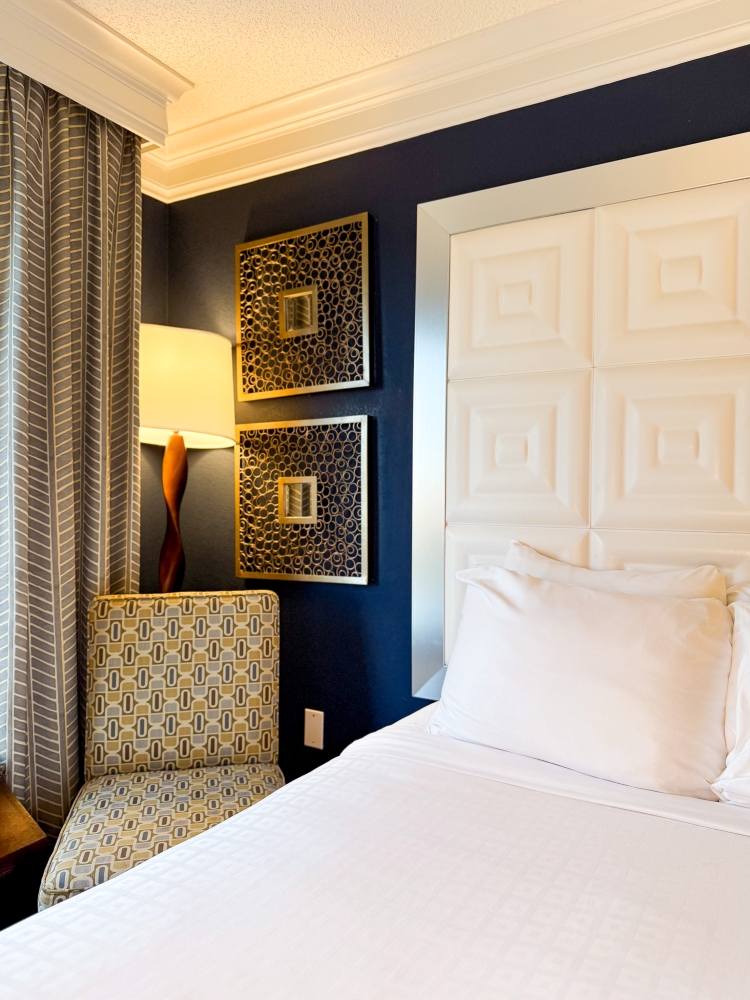 The side of a bed with a plush white headboard and navy walls, an armchair, and a floor lamp in a hotel room at Rosen Plaza.