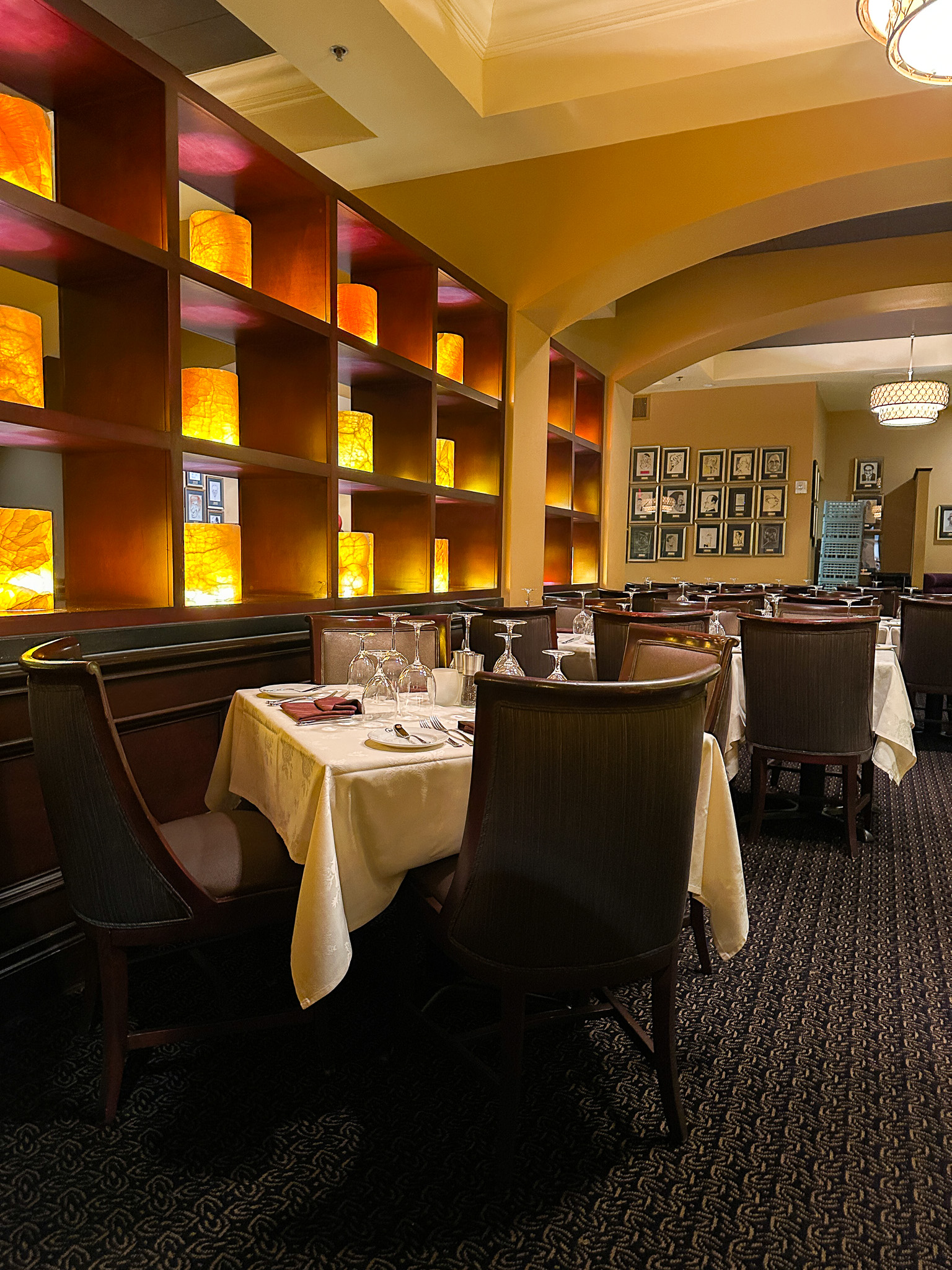 A dining room of a nice restaurant with tables adorned in white linens, leather chairs, and a wall of decorative boxes glowing with amber lights.
