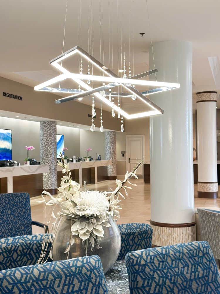 A hotel lobby with blue modern chairs, a contemporary chandelier, and a long marble front desk.