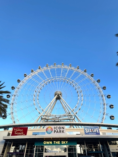 A large white Ferris wheel against a clear blue sky with several storefronts below.