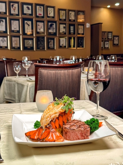 A lobster tail and steak on a white plate beside a glass of red wine on a restaurant table, surrounded by other tables and a wall full of art.