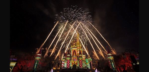Large streams of fireworks erupt against a dark sky over a whimsically lit Cinderella Castle.