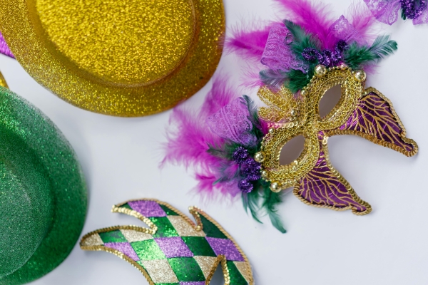 A sparkly, feathery mask sits on a white surface beside green and yellow glittery Mardi Gras hats. 