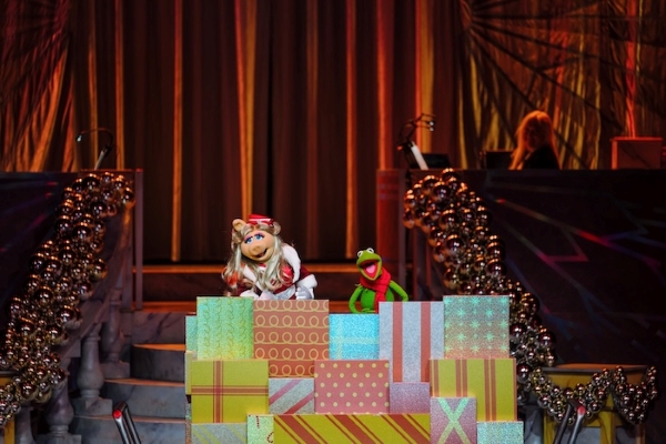 Miss Piggy and Kermit standing atop a pile of colorful presents in front of grand curtains onstage.