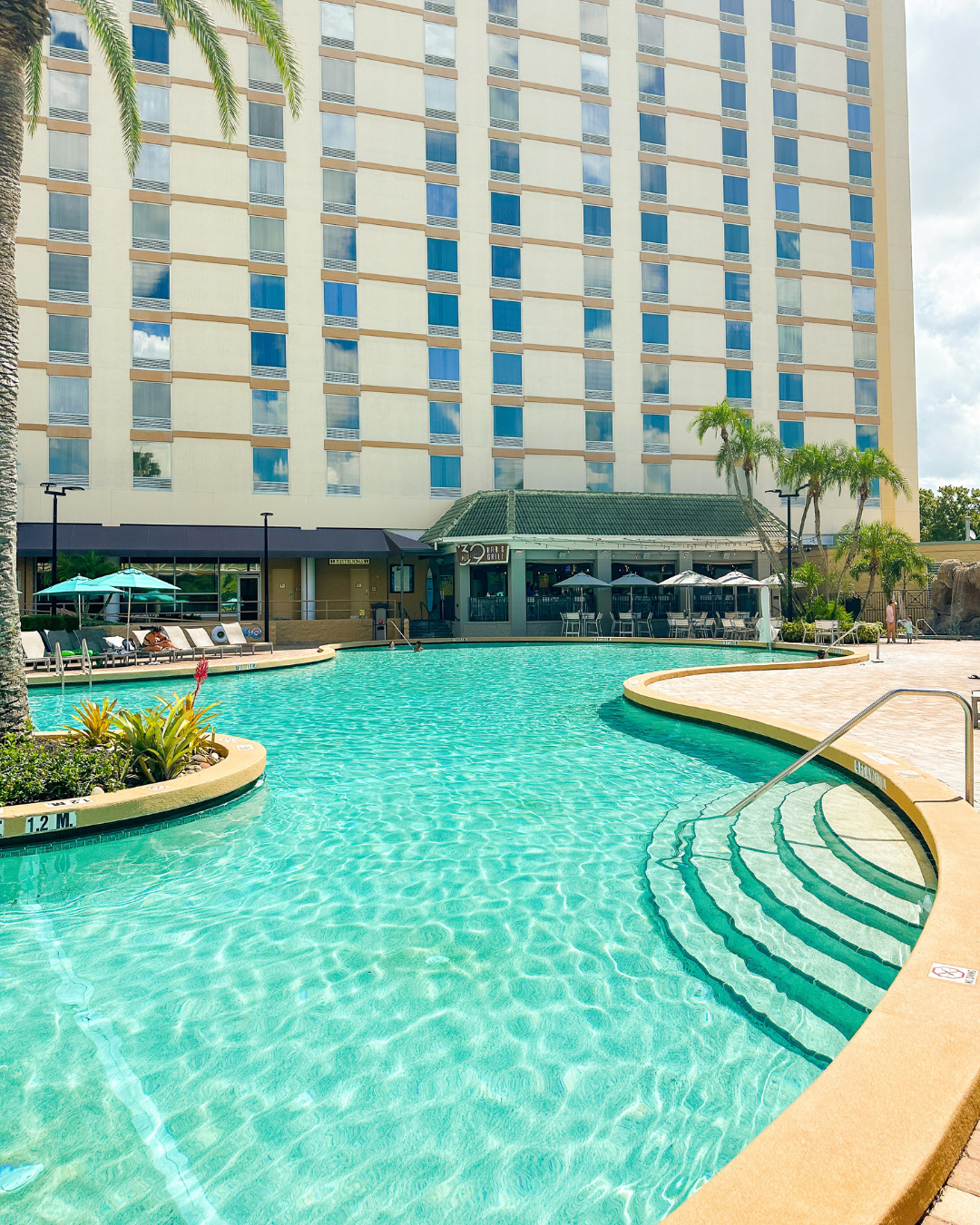 A curving clear blue pool surrounded by garden beds, blue umbrellas, and a tall building with a restaurant sign reading 39 Poolside Bar and Grill.