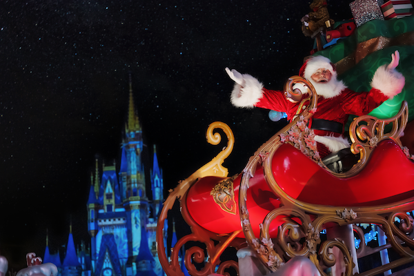 Santa Claus waving from a red sleigh with Cinderella Castle glowing blue at night in the background.
