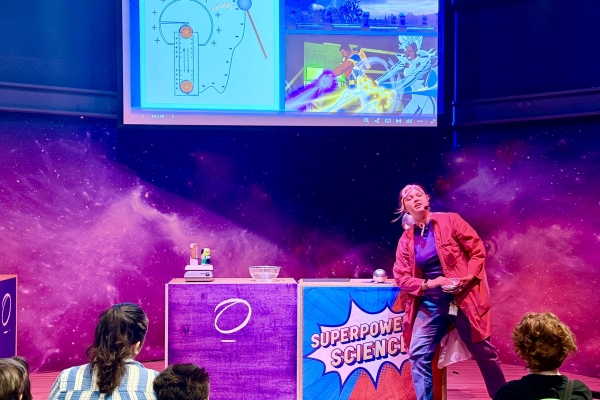 A woman performer in a red jacket stands on a stage in front of a screen and surrounded by fog during a science show.