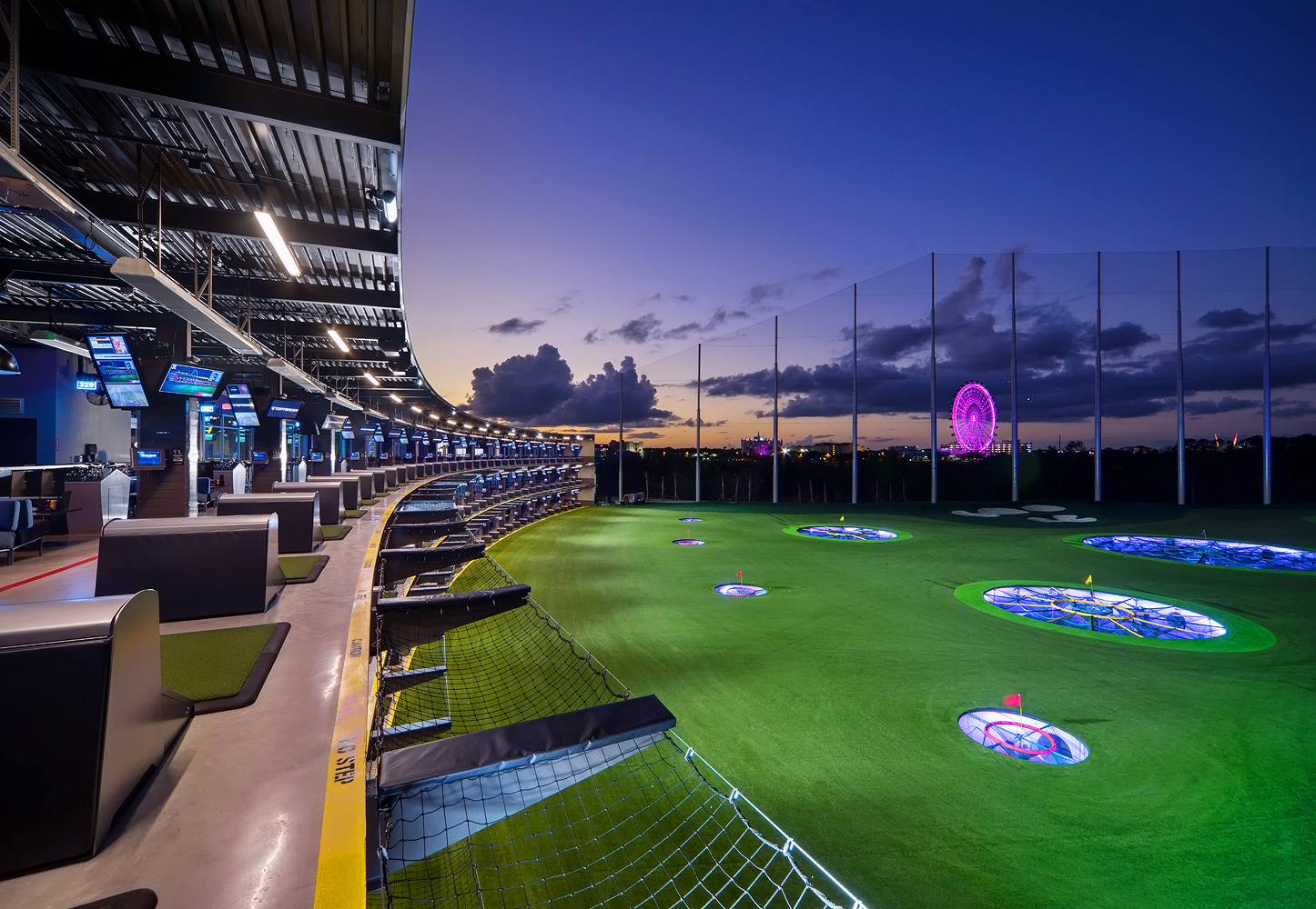 A curved row of golf bays at Top Golf overlooking a putting green with a pink glowing observation wheel in the background