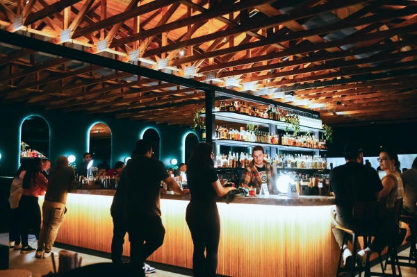 Silhouetted figures stand at a dark and moody bar with beams across the ceiling and a modern bar stocked with liquor bottles. 