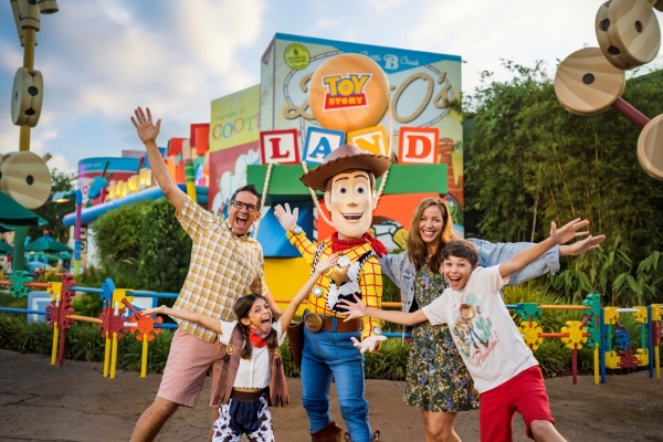 A family with a boy and a girl pose with Woody from Toy Story, arms outstretched, in front of Toy Story Land.