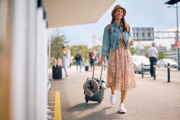 A woman in a dress drags her suitcase in front of an airport entrance with a phone in her hand.