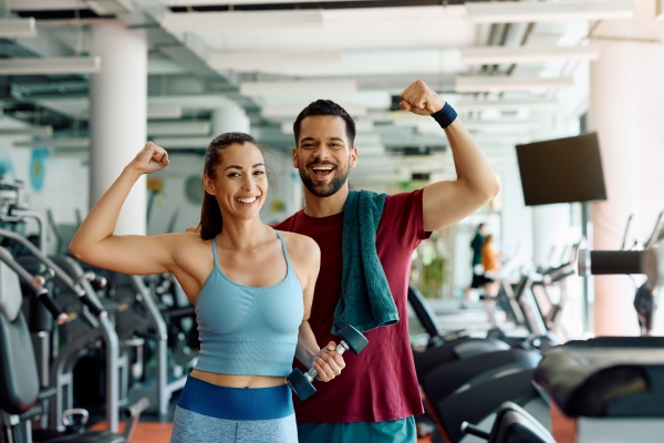 A man and a woman in workout gear flex their biceps standing in a gym facility, perhaps while staying fit on vacation.