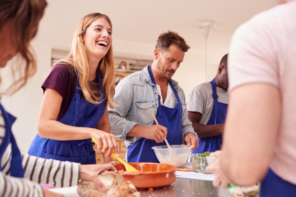 A group of adults wearing matching blue aprons smile as they cook together around a counter.