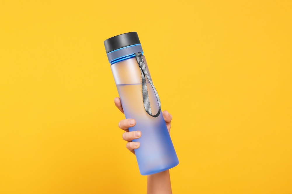 A hand holds a blue translucent water bottle against a yellow background.