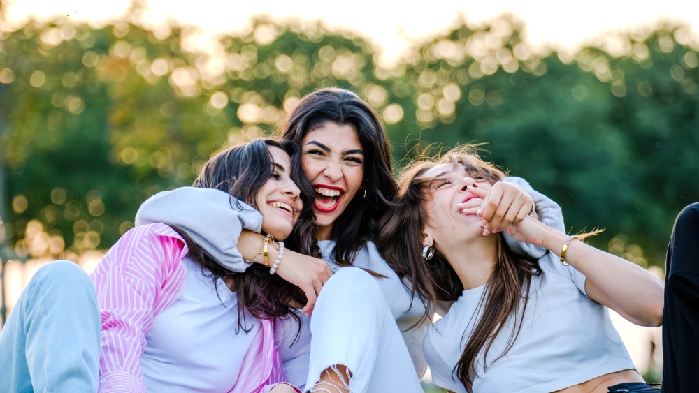 A woman sits with her arms playfully around two other women, giggling outdoors.
