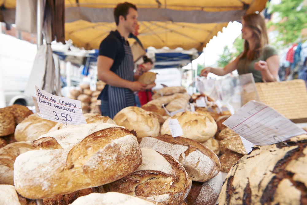 A man and woman stand across from one another under a tent, with a table of freshly baked bread between them.