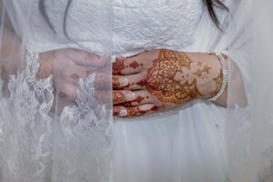 Close up shot of a bride in a white lace wedding gown and lace veil. Her hands, intricately decorated in henna, are folded over her midsection. 